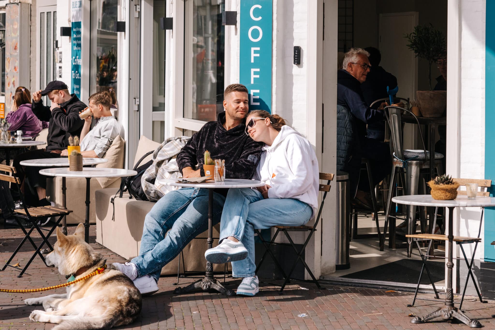 Een stel geniet van de zon op het terras van Blue Zone Espresso, met een hond in de voorgrond.