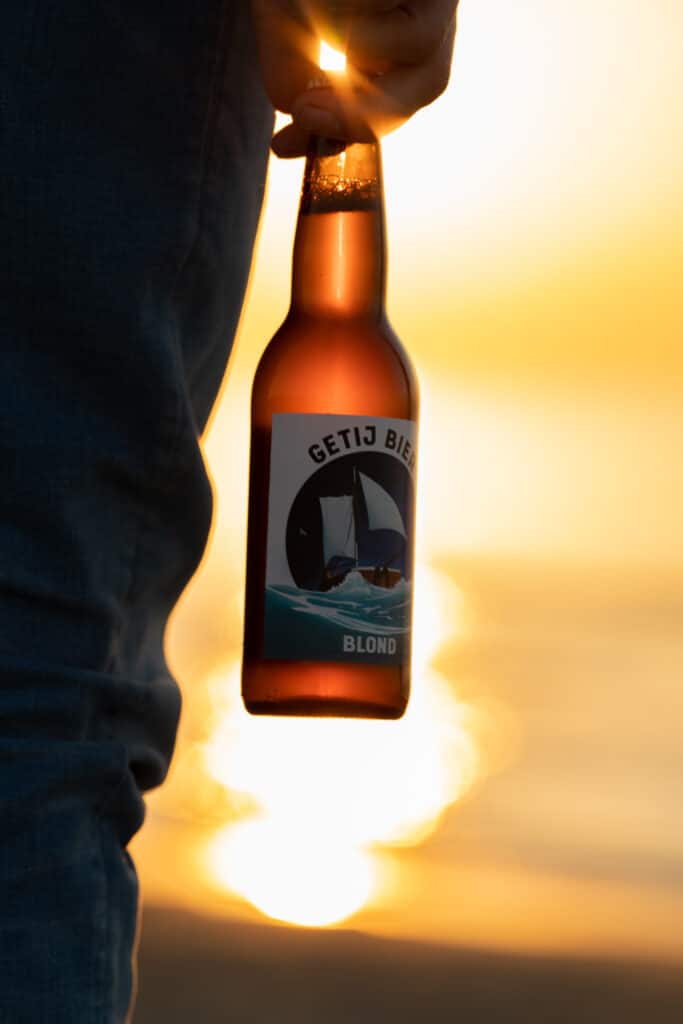 Iemand houdt een blond Getij bier vast met de zonsondergang op de achtergrond.